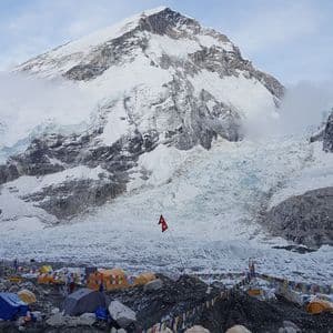 Un campamento base con tiendas de colores y banderas de oración se encuentra en una morrena rocosa debajo de una enorme montaña cubierta de nieve y un glaciar.