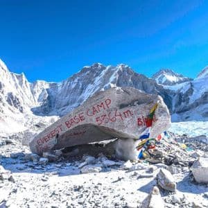 Der Markierungsfelsen des Everest-Basislagers, beschriftet mit seiner Höhe von 5364 m, befindet sich in einer verschneiten Landschaft, umgeben von Himalaya-Gipfeln unter klarem Himmel.