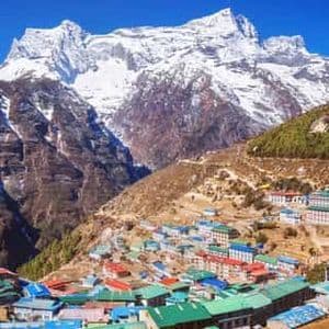 Un village aux toits colorés est niché à flanc de montagne escarpée, sous une grande montagne enneigée, sur fond de ciel bleu clair.