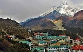 Un borgo dai tetti colorati è adagiato su un fianco roccioso, ai piedi di una grande montagna innevata, sotto un cielo nuvoloso.