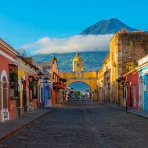 A cobblestone street lined with colorful colonial buildings leads to a yellow archway with a clock, with a volcano in the background under a blue sky.