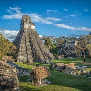An ancient stone pyramid and thatched-roof structures on a grassy clearing in the jungle under a partly cloudy blue sky.