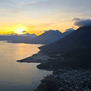 Vista elevada de un pueblo a orillas del lago rodeado de montañas al amanecer, con el resplandor del sol en el horizonte.