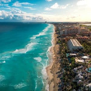 An aerial view of a crowded sandy beach with turquoise water, lined with hotels and palm trees under a blue sky with white clouds.