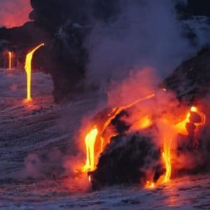 Molten lava pours from volcanic cliffs into the churning ocean, generating large clouds of steam and smoke at dusk.