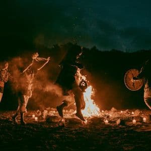 A group of people in traditional clothing performs a ritual dance around a large bonfire on the sand at night.