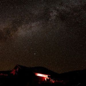 A vast, starry night sky with the Milky Way galaxy visible above a silhouetted house illuminated by red light.