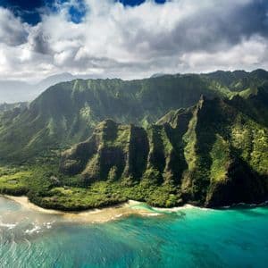 An aerial view of a dramatic, green mountain range descending into a turquoise ocean along a rugged coastline.