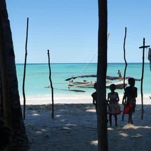Trois enfants en silhouette se tiennent sur une plage de sable, regardant un bateau sur la mer turquoise sous un ciel bleu clair.