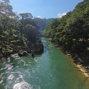 Un río turquesa fluye a través de un valle con exuberantes colinas verdes a ambos lados, bajo un cielo azul soleado.