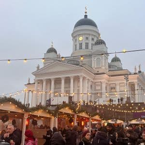 Un vivace mercatino di Natale con folla attorno a bancarelle in legno, di fronte a una grande cattedrale dalla cupola bianca sotto un cielo coperto.