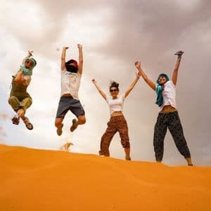 Un gruppo WeRoad di quattro persone che saltano insieme con le braccia alzate in cima a una duna di sabbia arancione.