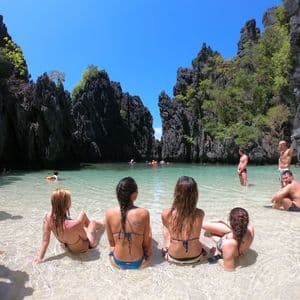 A WeRoad group trip relaxing in the clear shallow water of a lagoon surrounded by tall, dark rock formations under a sunny sky.
