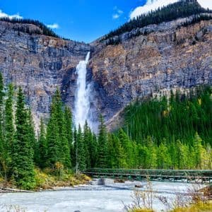 Una cascata imponente scende da una parete rocciosa stratificata in un fiume impetuoso, con un ponte verde e una pineta in primo piano.