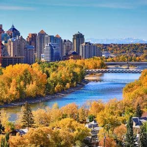 A city skyline overlooks a winding river, its banks lined with golden autumn trees, with distant mountains under a blue sky.