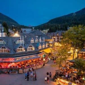 Una vivace piazza del paese al tramonto, con persone che cenano nei ristoranti all'aperto e passeggiano tra edifici illuminati in modo caldo ai piedi delle montagne.