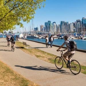 Ciclisti e pedoni percorrono un sentiero lastricato lungo un porto turistico pieno di barche a vela, con lo skyline di una città sullo sfondo in una giornata di sole.