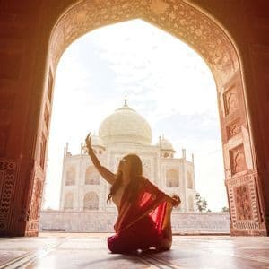 Una donna in un sari rosso pratica yoga sotto un arco di pietra, che incornicia la vista di un mausoleo a cupola di marmo bianco.