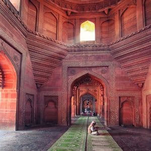 Un uomo è seduto su un lungo tappeto verde in una grande sala di arenaria rossa, circondato da archi finemente intagliati.