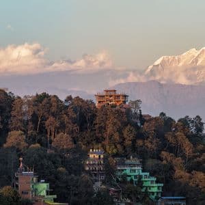 Veduta panoramica di un paese su una collina boscosa con grandi montagne innevate sullo sfondo all'alba.