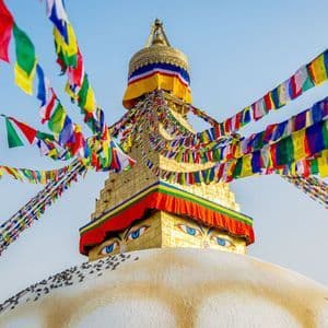 Uno stupa dorato con grandi occhi blu dipinti è adornato da colorate bandiere di preghiera che si stagliano contro un cielo azzurro e limpido.