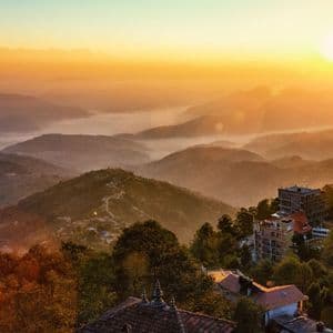Un'alba dorata illumina montagne e valli nebbiose e stratificate, immerse nella nebbia, con edifici annidati tra gli alberi in primo piano.