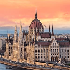 Ein großes, verziertes Regierungsgebäude steht an einem Flussufer in einer Stadt während eines lebhaften Sonnenuntergangs.