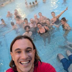 Un hombre se hace una selfie con su grupo de viaje WeRoad, sonriendo y relajándose en un concurrido baño termal.