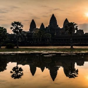 Un templo antiguo con varias torres se recorta contra el amanecer y se refleja en un lago con palmeras.