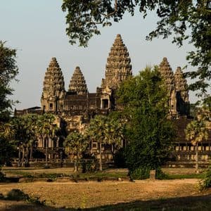 Antiguo templo de piedra con múltiples torres ornamentadas, enmarcado por exuberantes árboles y palmeras bajo un cielo despejado.