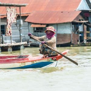 Una persona con sombrero de paja y mascarilla rema en una barca pequeña en un pueblo flotante con casas sobre pilotes.