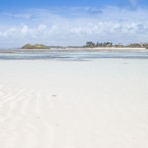 Una vasta distesa di sabbia bianca con increspature è visibile sotto acqua bassa e limpida, con una costa tropicale in lontananza sotto un cielo azzurro.