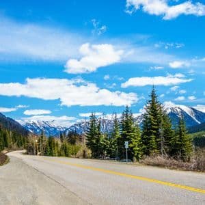 Una strada deserta si snoda attraverso una valle boscosa verso distanti montagne innevate sotto un cielo azzurro brillante con nuvole.