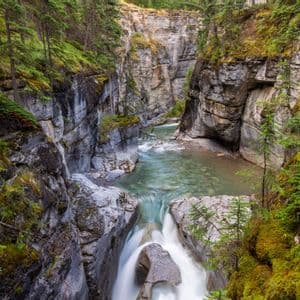 Una cascata si getta in un fiume che scorre in un profondo canyon roccioso, fiancheggiato da muschio e pini.