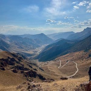 Una vista panoramica di una vasta valle di montagna con una strada tortuosa e un fiume che serpeggia attraverso il paesaggio arido sotto un cielo blu.