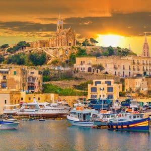 Un porto pieno di barche si trova di fronte a una città collinare con una chiesa imponente, il tutto immerso nella luce di un tramonto dorato.