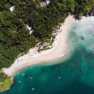 Una vista aerea dall'alto di una spiaggia tropicale con sabbia bianca, delimitata da una rigogliosa foresta verde e acqua turchese cristallina con diverse imbarcazioni.