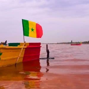 Un barco colorido con la bandera de Senegal flota en aguas de color rosa rojizo bajo un cielo nublado.