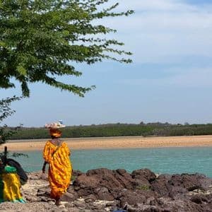 Una mujer con un vibrante vestido naranja camina por una costa rocosa, equilibrando una bandeja de productos en su cabeza junto a aguas turquesas.