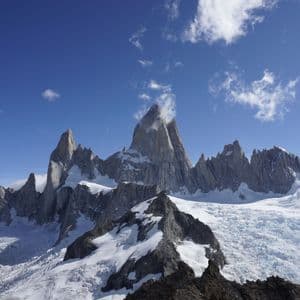 A range of jagged, rocky mountains covered in snow and glaciers under a bright blue sky with sparse clouds.