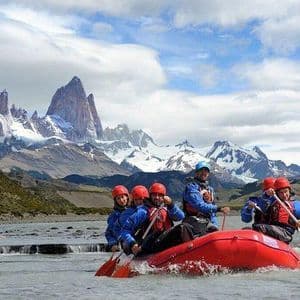 Un voyage de groupe WeRoad en rafting dans un radeau rouge sur une rivière, avec une chaîne de montagnes enneigées en arrière-plan.