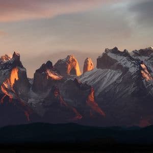 Ein Panoramablick auf eine schneebedeckte Bergkette mit zerklüfteten Gipfeln, beleuchtet vom warmen Licht des Sonnenaufgangs oder Sonnenuntergangs.