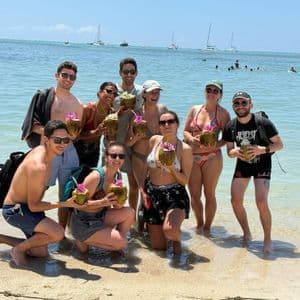 Un groupe WeRoad sourit et pose avec des boissons à la noix de coco, les pieds dans l'eau peu profonde d'une plage.