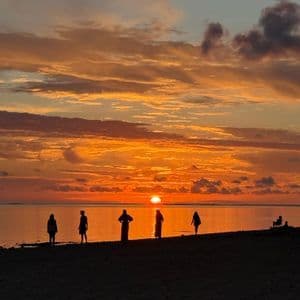 Silhouettes d'un groupe WeRoad en voyage, debout sur une plage, observant un coucher de soleil orange sur l'eau.