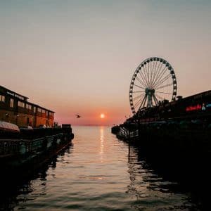 Una grande ruota panoramica ed edifici su un molo si stagliano contro il sole al tramonto, che si specchia sull'acqua sottostante.