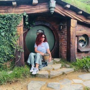 A woman with curly red hair sits on the stone steps of a hobbit-style house, holding a small wooden bucket.