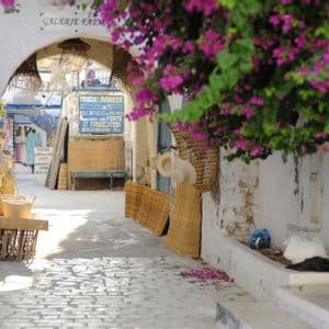 Eine sonnenbeschienene Kopfsteinpflastergasse auf einem Markt, mit Kunsthandwerk zum Verkauf und leuchtend rosa Bougainvillea-Blüten, die darüber hängen.