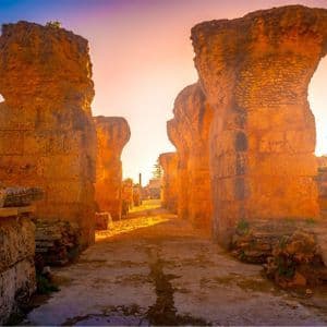 Ein Pfad schlängelt sich durch antike Steinruinen mit imposanten Säulen, beleuchtet vom goldenen Licht einer untergehenden Sonne.