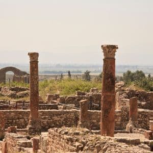 Antike Steinruinen mit verwitterten korinthischen Säulen und niedrigen Mauern, die eine dunstige grüne Landschaft überblicken.