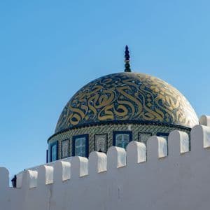 Eine reich verzierte goldene und blaue Kuppel einer Moschee ragt über einer weißen zinnenbewehrten Mauer vor einem klaren blauen Himmel empor.